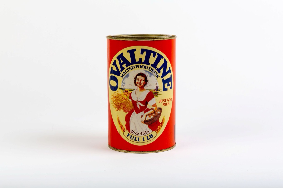A red and cream Ovaltine tin, sixteen ounces, photographed on a plain white background, showing the Ovaltine Girl with a sheaf of wheat and a basket of eggs.