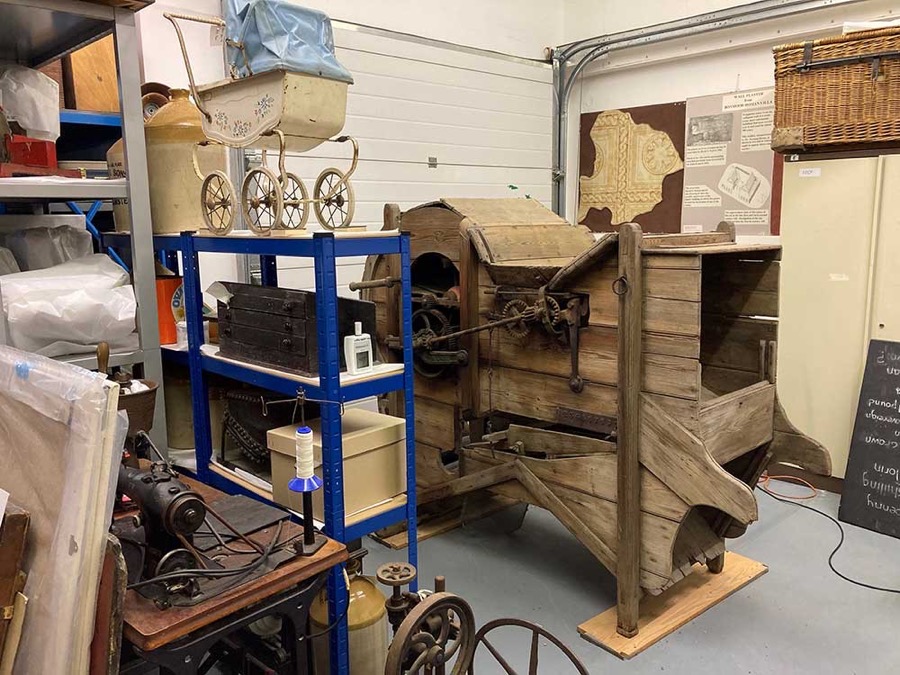 Inside the Museum Store at Clarence Road: blue steel racking holding a Victorian pram, stoneware jars and boxed objects, beside a large wooden threshing machine.